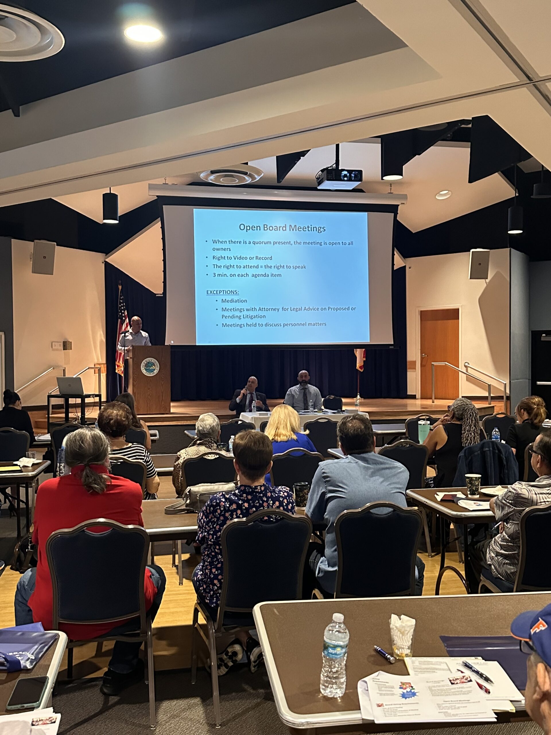 A large room in Delray Beach is filled with people seated at tables facing a stage. A live presentation is projected on a screen at the front. A speaker stands at a podium, and two individuals are seated on stage. Attendees are focused on the presentation.