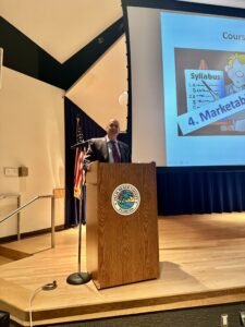 At a wooden podium on stage, attorney Barry Ansbacher is delivering a LIVE presentation. The screen lights up the room as audiences in Delray Beach gather to tune into this engaging session.