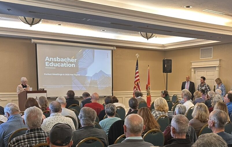A group of people seated in a conference room, facing a speaker at a podium. A presentation screen displays "Ansbacher Education." The room is well-lit with chandeliers, and the American flag is visible.