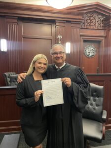 A smiling woman in a suit stands next to a judge in a courtroom, both holding an official document. The judge wears a black robe, and the room features dark wood paneling and a judge’s bench in the background.