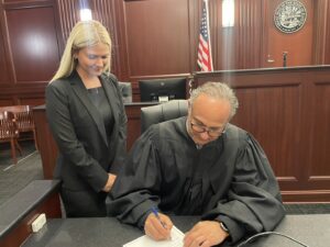 A judge in a black robe signs a document at a desk in a courtroom, while a woman in a black suit stands beside him, smiling. An American flag and a seal are visible in the background.