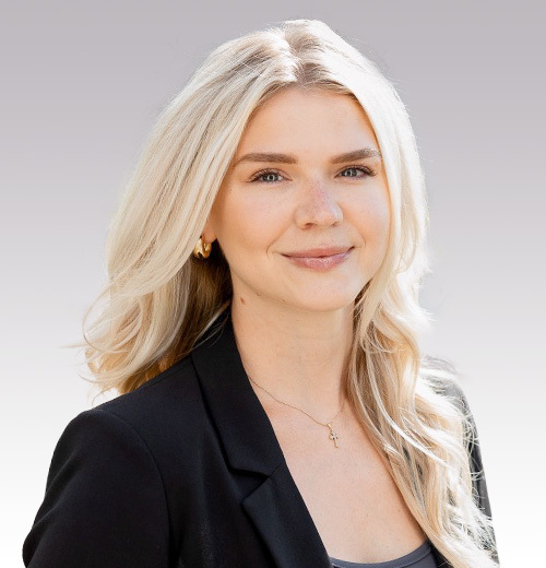 Brooke M. Cleary, a woman with long, wavy blonde hair, wears a black blazer, gray top, and gold necklace, smiling softly against a light gray background.