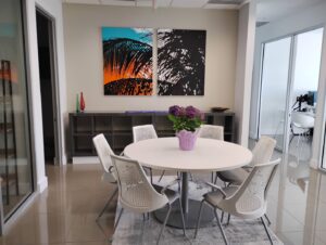 A modern office meeting room at Ansbacher Law in Coral Gables features a round white table, six white chairs, a vibrant purple potted plant centerpiece, abstract wall art, and glass walls leading to other office spaces.