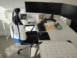 A modern office desk with three monitors, a keyboard, mouse, phone, calendar, and stationery. A white and blue tote bag hangs on the black chair. Sunlight shines on the glossy floor—perfect for any Coral Gables or Ansbacher Law professional.