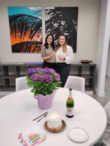 Two women stand smiling behind a table with a small cake, sparkling cider, a potted purple hydrangea, party napkins, and plates. Colorful artwork hangs on the wall behind them at what seems to be an Ansbacher Law celebration in Coral Gables.