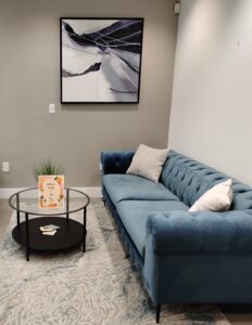 A modern living room in Coral Gables features a blue tufted sofa, white and gray cushions, an abstract painting, and a round glass coffee table with a plant, cards, and a book set on a patterned rug—an inviting space for Ansbacher Law clients.