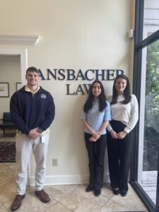 Three people, including the 2025 Summer Clerks, stand indoors smiling in front of a cream-colored wall with blue letters that read "ANSBACHER LAW." Sunlight streams in through large windows on the right.