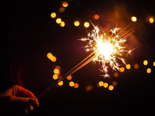 A hand holds a lit sparkler against a dark background with glowing golden bokeh lights, capturing the festive spirit of the Fourth of July in Florida.