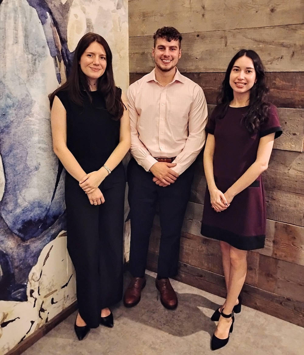 Three young adults stand side by side indoors, dressed in business casual attire, smiling at the camera. Behind them is a wooden wall and a colorful, abstract mural.