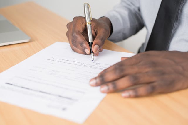 A person in business attire signs a Real Estate Purchase Agreement on a wooden desk with a silver and gold pen. A laptop is partially visible in the background.