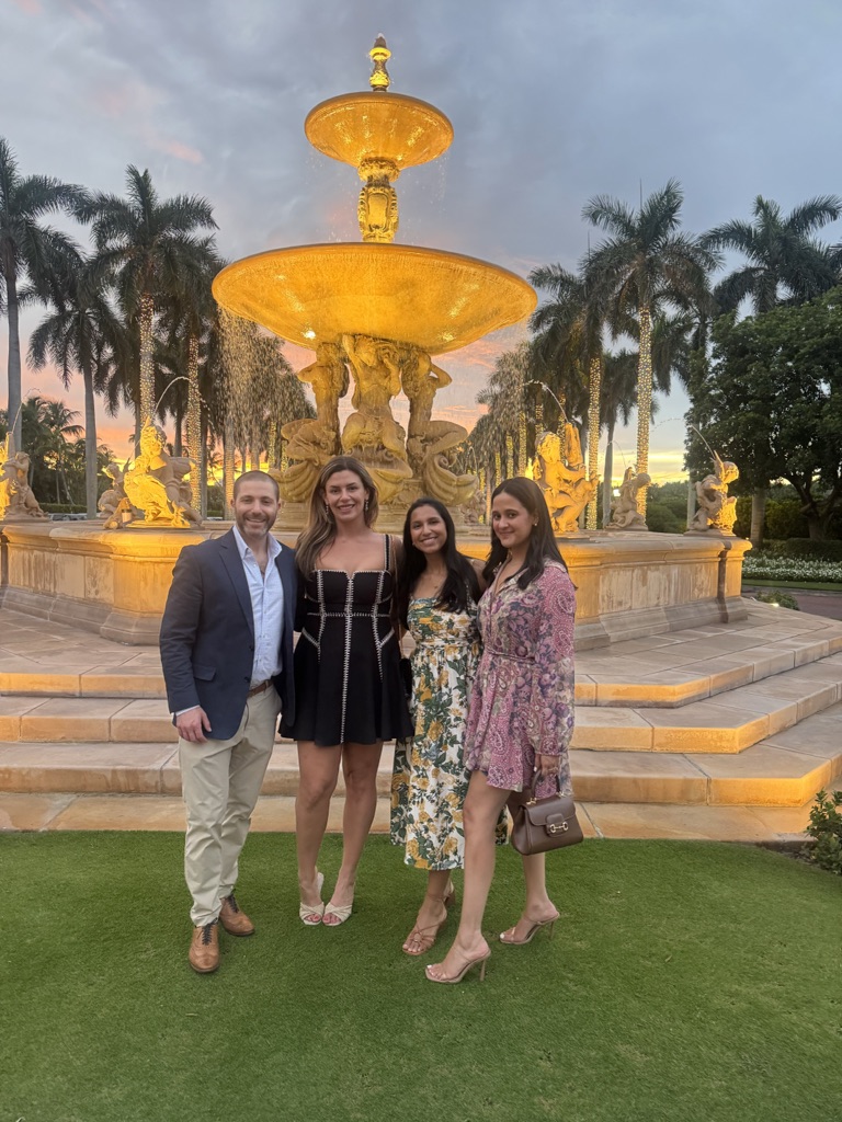Four people pose and smile in front of a large, ornate, illuminated fountain surrounded by palm trees at sunset, perhaps celebrating a milestone in trust law or real property achievements. Three women wear dresses and sandals; one man wears a suit jacket.