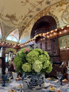 A large bouquet of green hydrangeas in a blue and white vase sits on an elegant dining table at Flagler College, inside a grand, ornately decorated hall with detailed murals and stained glass windows.