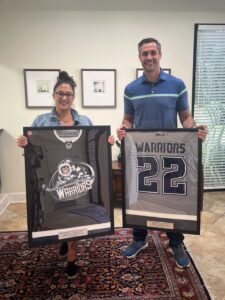 Two people stand indoors on a patterned rug, each smiling and holding framed "Warriors" jerseys. The woman’s jersey features a tiger logo; the man's displays the number 22. Thank You Jacksonville Icemen Warriors for honoring their achievements!.