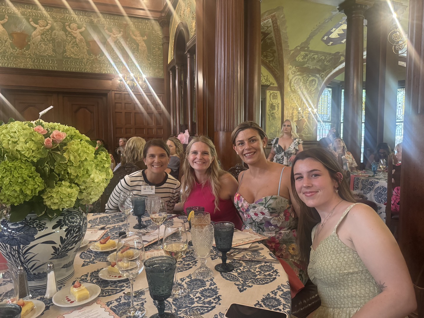 Four women sit together at a table set with plates, glasses, and a floral centerpiece in an ornate room at Flagler College, smiling at the camera during the Power of the Purse event presented by Ansbacher Law.