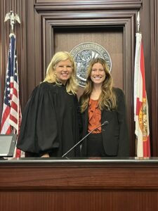 Two women stand and smile in a courtroom. One wears a black judge’s robe, and the other, Attorney Kelly Welch of Ansbacher Law, is in business attire. Behind them are the U.S. and Florida flags, with a large seal on the wooden wall.