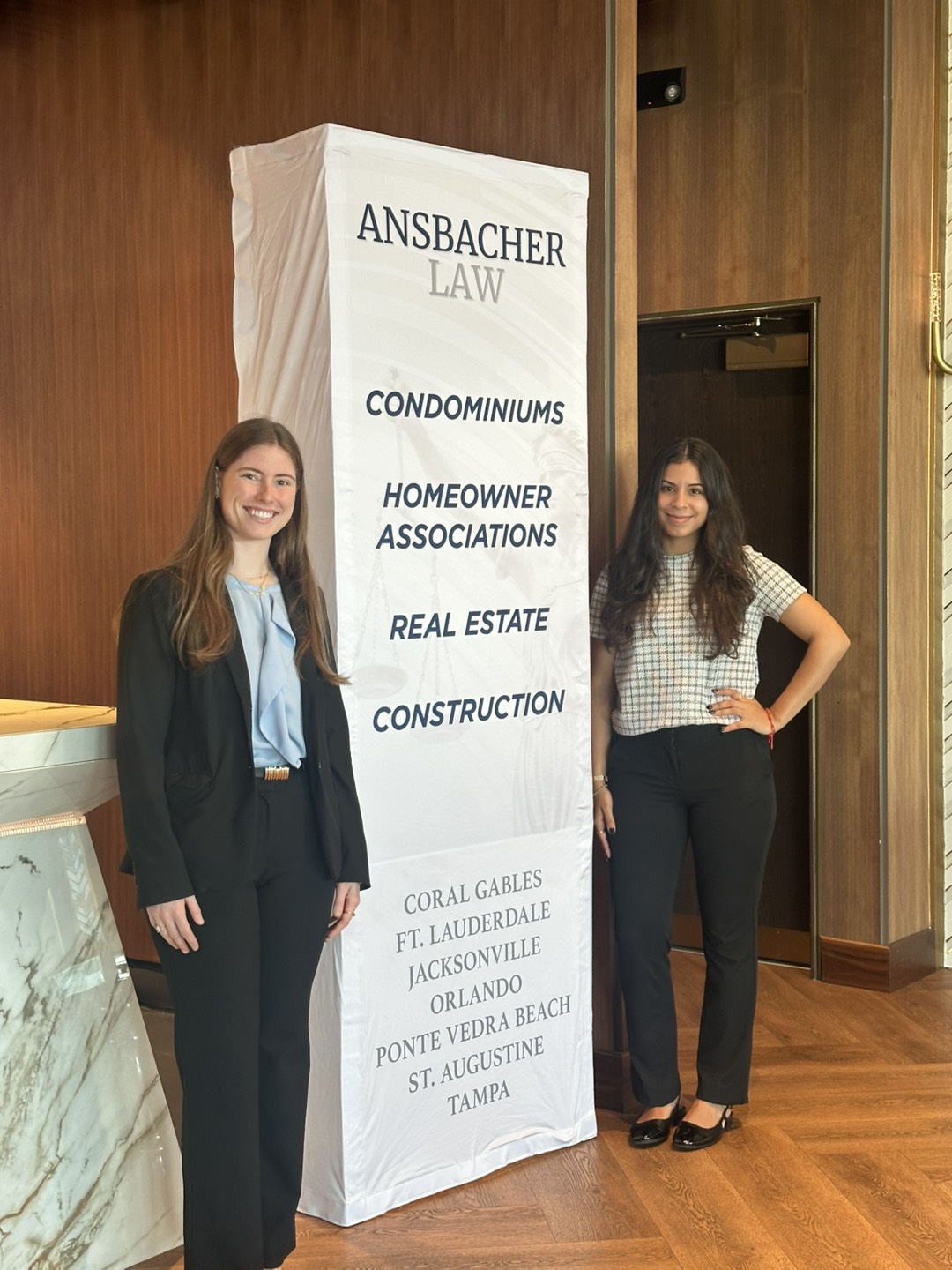 Two women in business attire stand smiling next to a tall Ansbacher Law sign, highlighting services like condominiums, SIRS, Milestones, homeowner associations, real estate, and construction, as well as Brunch and Learn events across Florida cities.