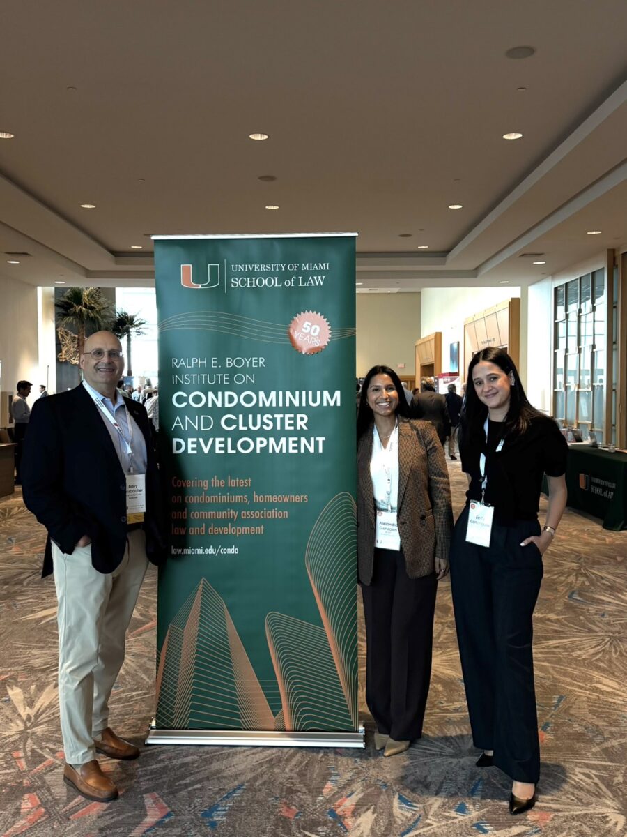 Three people stand smiling beside a vertical banner for the University of Miami School of Law’s Boyer Institute, celebrating 50 years of the Ralph E. Boyer Institute on Condominium and Cluster Development inside a large, well-lit conference space.