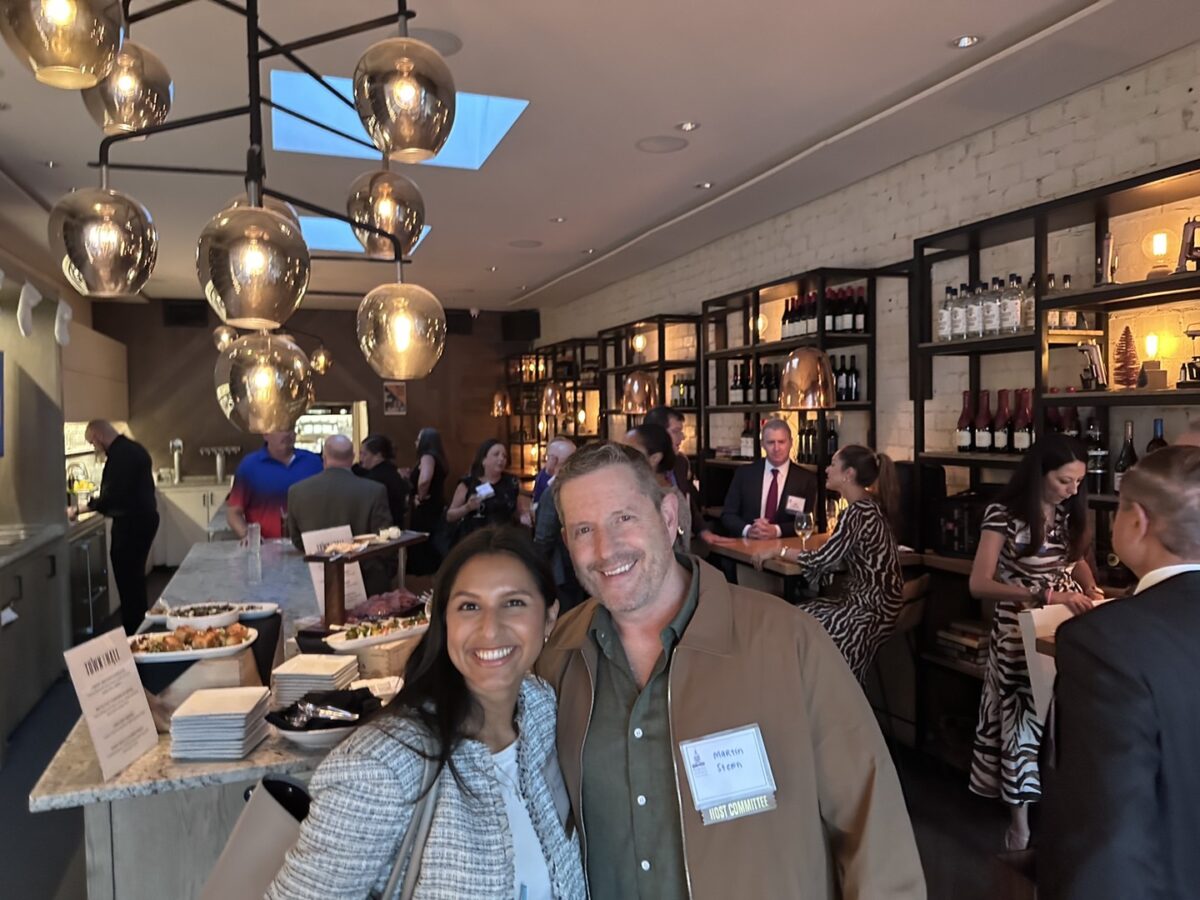 A smiling woman and man pose together at a JALA Event in a stylish bar or restaurant. Other guests, including members of Ansbacher Law, mingle near shelves with bottles and snacks on the counter. Warm lighting creates a cozy atmosphere.