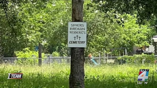 A sign reading "SPIERS BROWN ROBERTS CEMETERY" with two cross symbols is attached to a tree in a grassy area, with trees and a wire fence in the background. The News4JAX logo appears in the bottom corners.