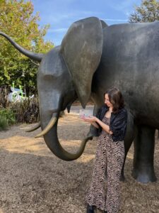 A woman in patterned pants and a black top stands beside a large elephant statue outdoors, smiling and presenting as if celebrating Best Places to Work 2025, with lush greenery in the background—a proud moment for Ansbacher Law.