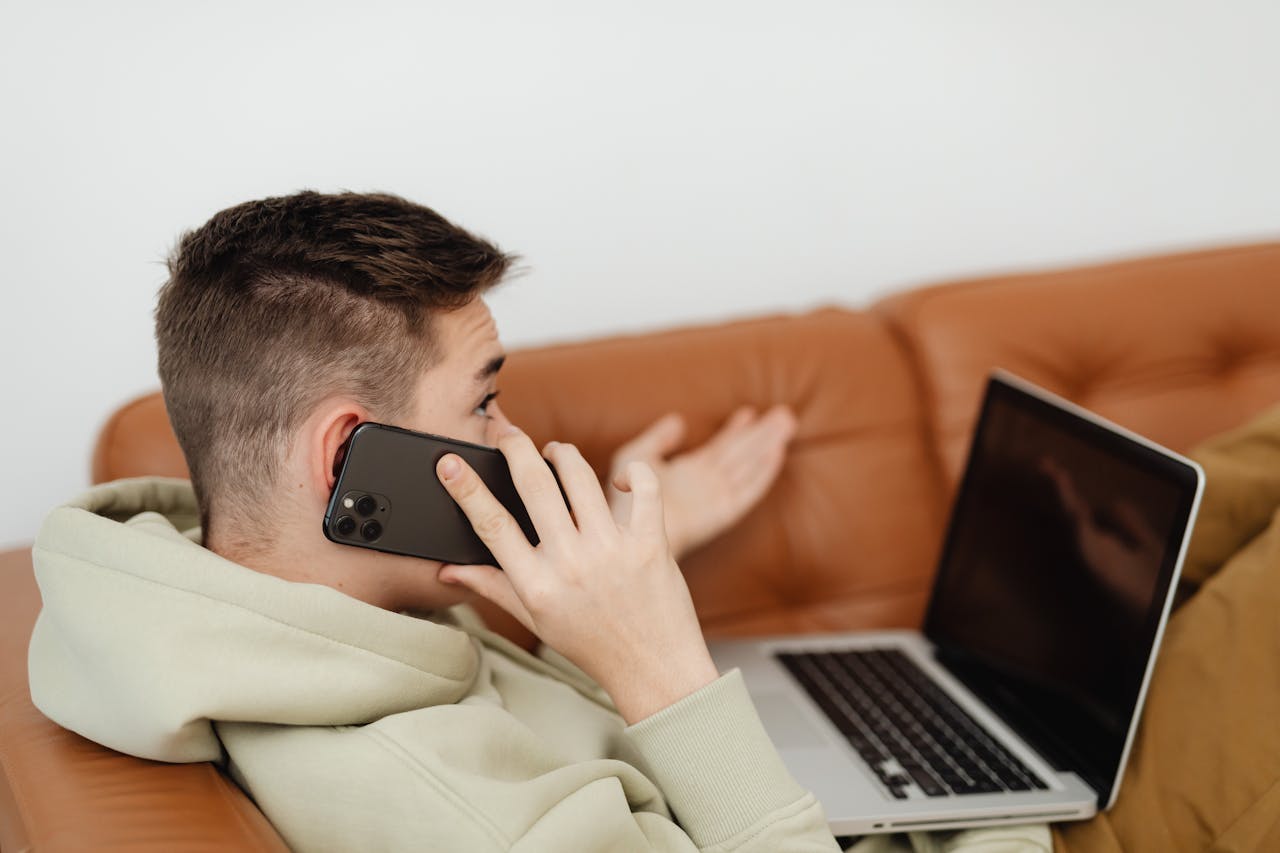 A person sits on a brown couch, holding a smartphone to their ear—possibly talking to an insurance adjuster—while gesturing with one hand and looking at an open laptop on their lap, carefully considering what not to say.