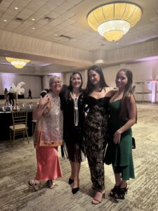 Four women dressed in formal evening attire stand together and smile at the Gala & Awards Dinner hosted by CAI Southeast Florida, with elegant chandeliers and round tables illuminating the grand ballroom.