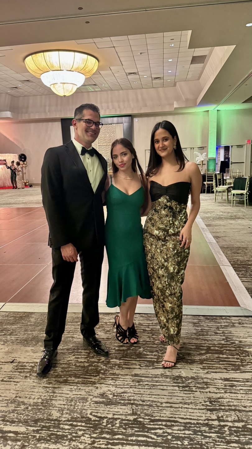 Three people dressed formally stand together at the Gala & Awards Dinner. The man on the left wears a black tuxedo, the woman in green is in the middle, and the woman on the right dons a strapless black and gold dress.