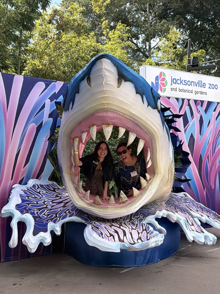 Two people smile while posing inside a large, colorful shark mouth display at the Jacksonville Zoo and Gardens, one of 2025’s Best Places to Work, surrounded by vibrant decorations and greenery in the background.