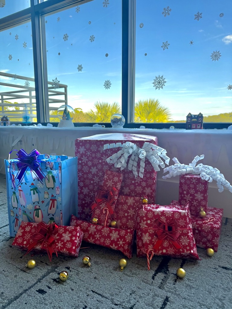 Colorful wrapped presents with bows sit on a carpet in front of a large window decorated with snowflake stickers. Part of the 2025 Holiday Project by Ansbacher Law, this joyful scene captures the spirit of JBA Dear Santa.