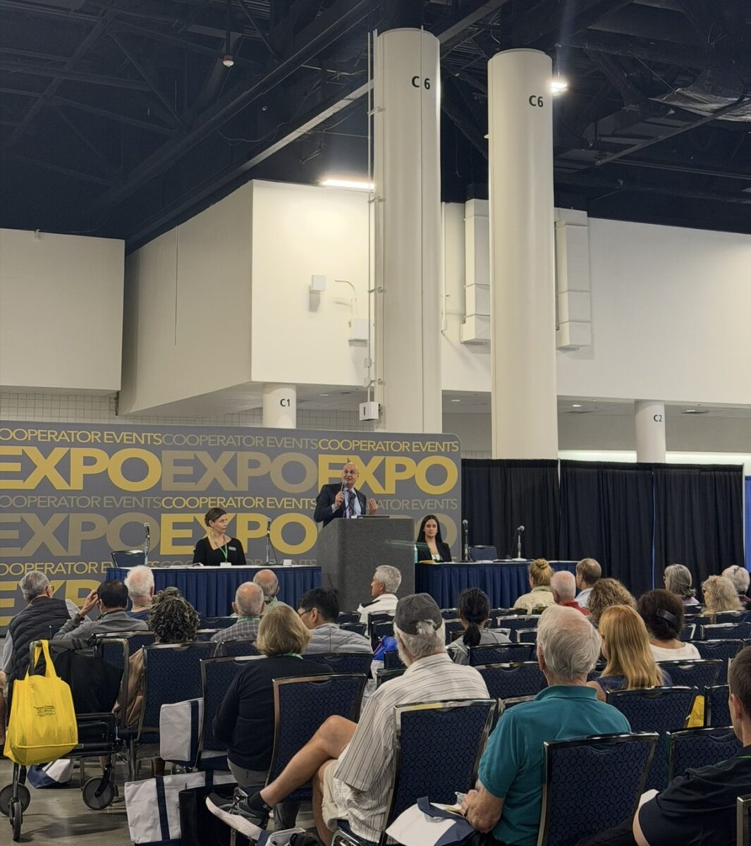 A speaker stands at a podium addressing an audience seated in rows at a South Florida expo event. Two panelists are seated at a table on stage, and large “EXPO” banners with the Cooperator Events logo are displayed in the background.