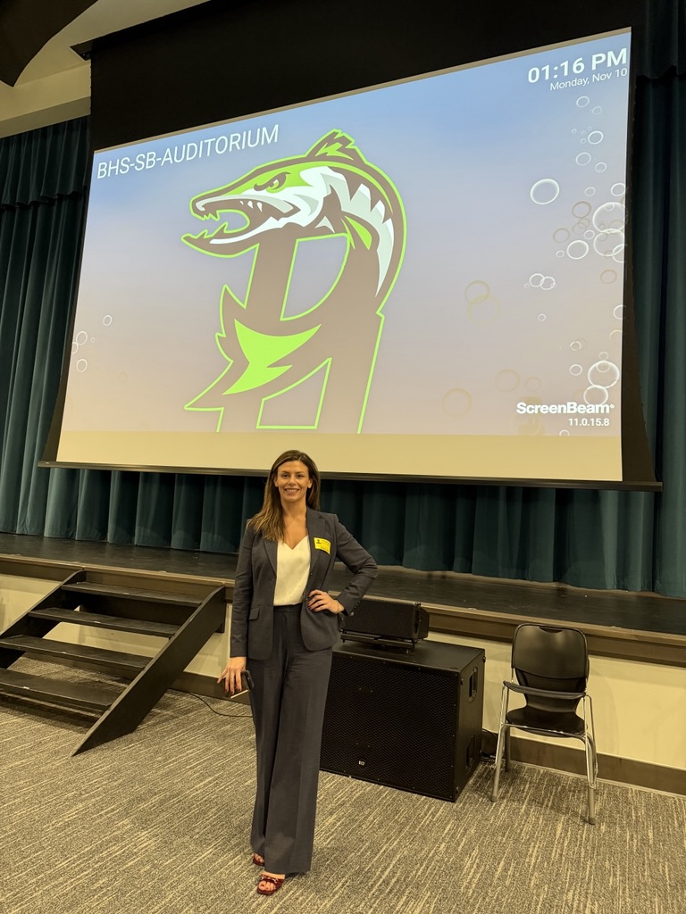 Hannah Rullo, a woman in a gray suit, stands smiling in front of a stage at Beachside Highschool. Behind her, the screen shows a green and yellow dragon logo with "BHS-SB-AUDITORIUM" and the time—01:16 PM, Monday, Nov 10—highlighting her teaching event.
