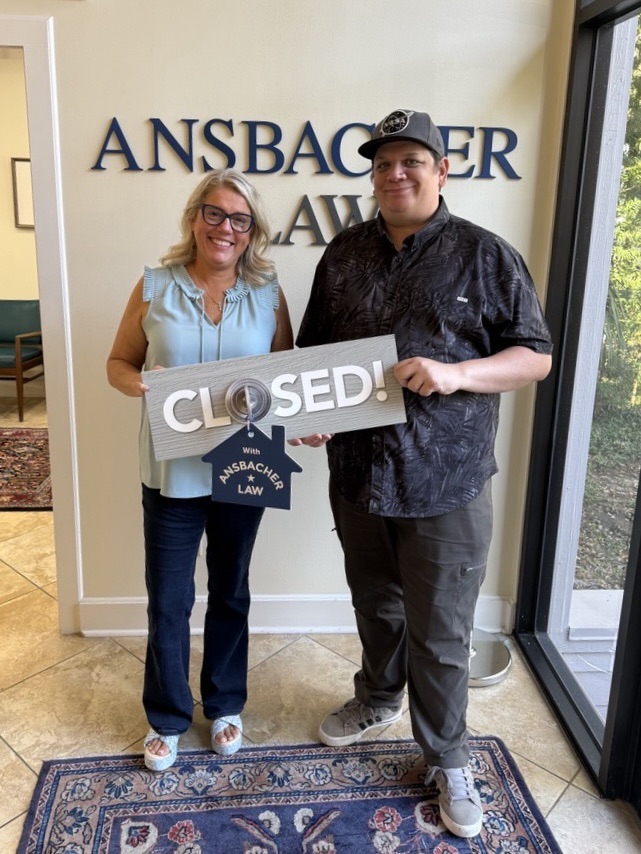 Two people stand smiling indoors, holding a large sign that says “CLOSED!” with a small tag reading “with Ansbacher Law,” celebrating a successful closing. The Ansbacher Law logo is visible on the wall behind them.