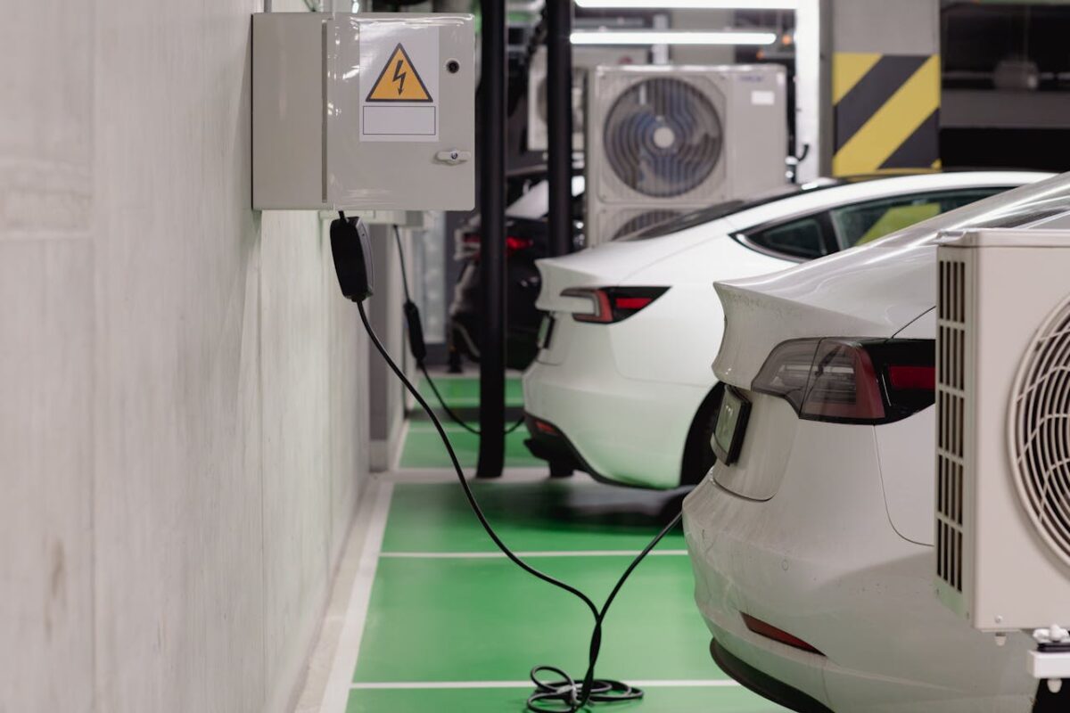 Two white electric vehicles are parked in a garage of a Florida condominium, with one plugged into a wall-mounted charging station. Owners and boards have added air conditioning units and safety markings along the walls.