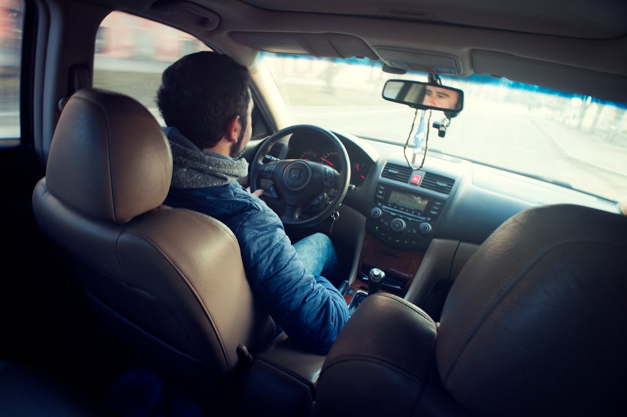 A man wearing a blue jacket is driving a car, viewed from the backseat. The dashboard and steering wheel are visible, and the empty road ahead highlights safe driving—helping avoid common types of car accidents in Florida.