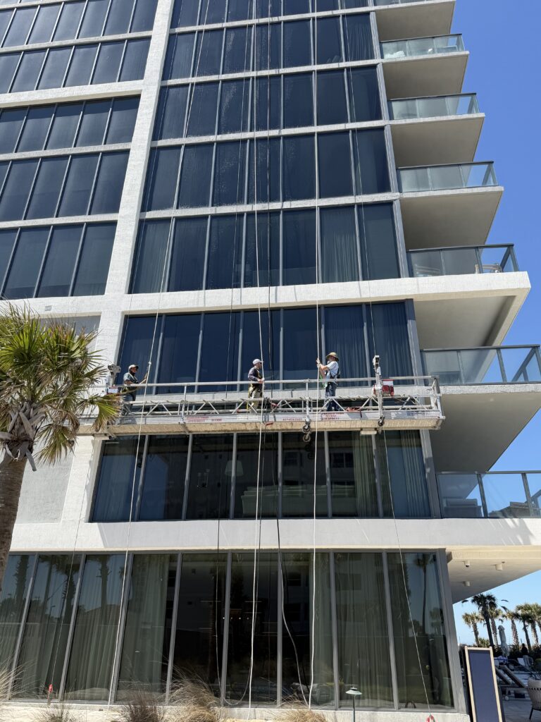 Four workers on a suspended platform clean windows on the exterior of a modern, multi-story glass building. Ropes hang down the facade, and balconies and clear blue sky are visible.