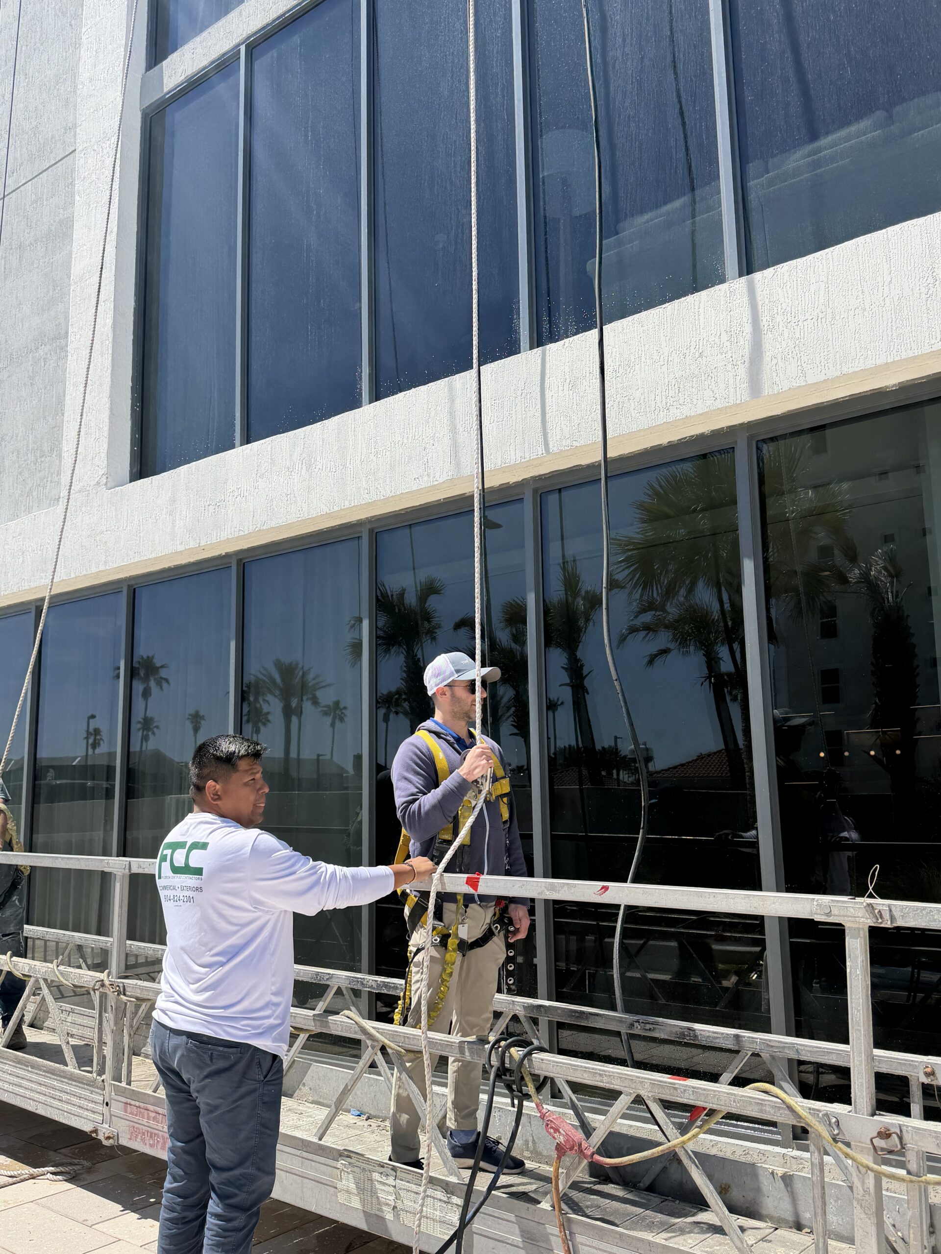Two workers are in action on a suspended platform outside a tall building with large glass windows. One adjusts harness equipment while the other holds a safety rope. Reflections of palm trees shimmer in the windows, highlighting their work on site.