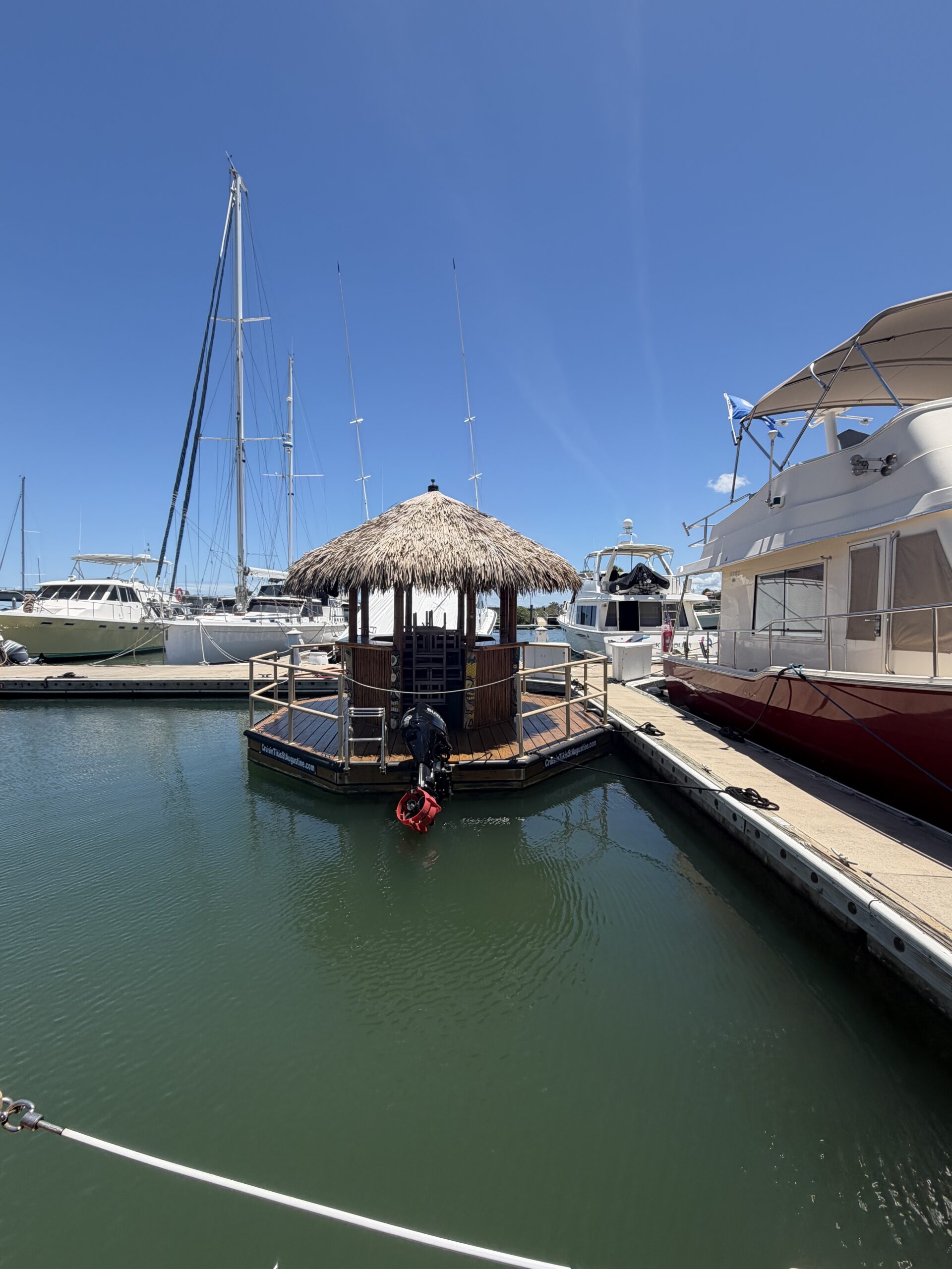 A floating tiki hut with a thatched roof is docked in a Florida marina, surrounded by boats and yachts under a clear blue sky. A person sits near the edge of the hut by the water, enjoying the view near the Ansbacher Law office.