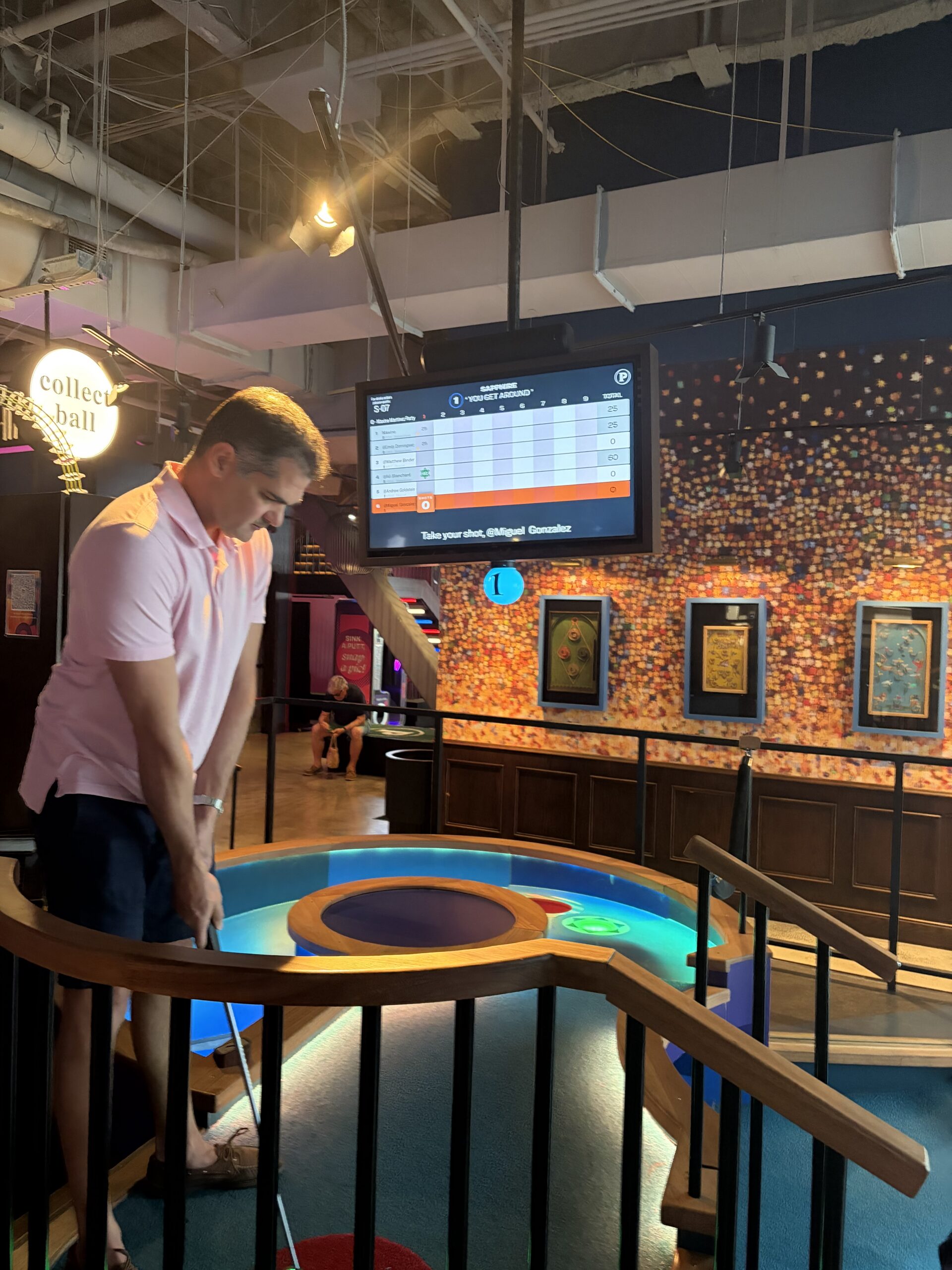 A man in a pink shirt lines up a putt at an indoor mini-golf course, focused on the ball. A large electronic scoreboard and colorful wall art decorate the background, giving this Florida office some playful charm.
