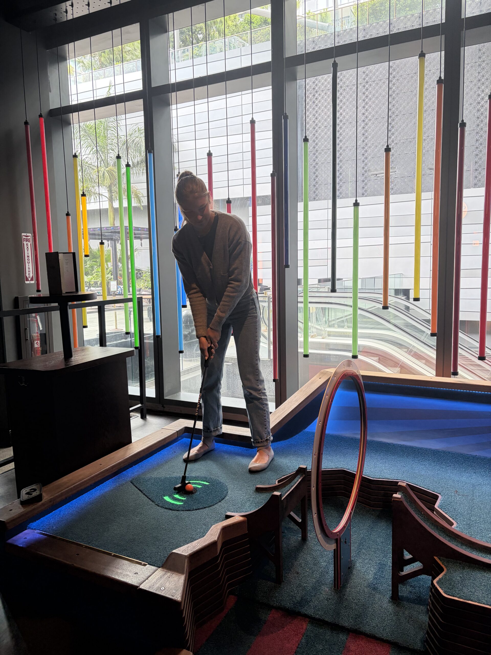 A person plays mini-golf indoors at an office in Florida, aiming to hit a golf ball through a red hoop obstacle. Colorful vertical pipes hang by the window behind them as sunlight streams in, creating a lively scene.
