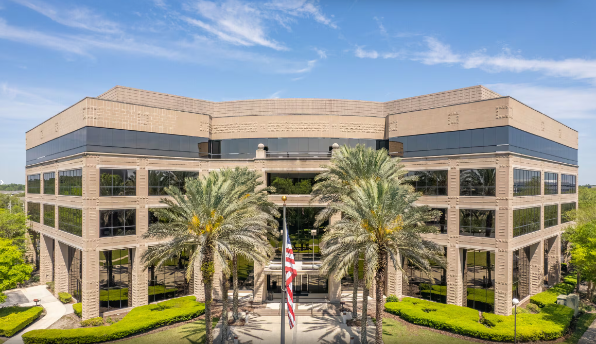 A large, modern office building with reflective windows and a tan stone exterior stands symmetrically amid palm trees and greenery. The U.S. flag waves in front under a clear blue sky—a prime location for Ansbacher moving services.