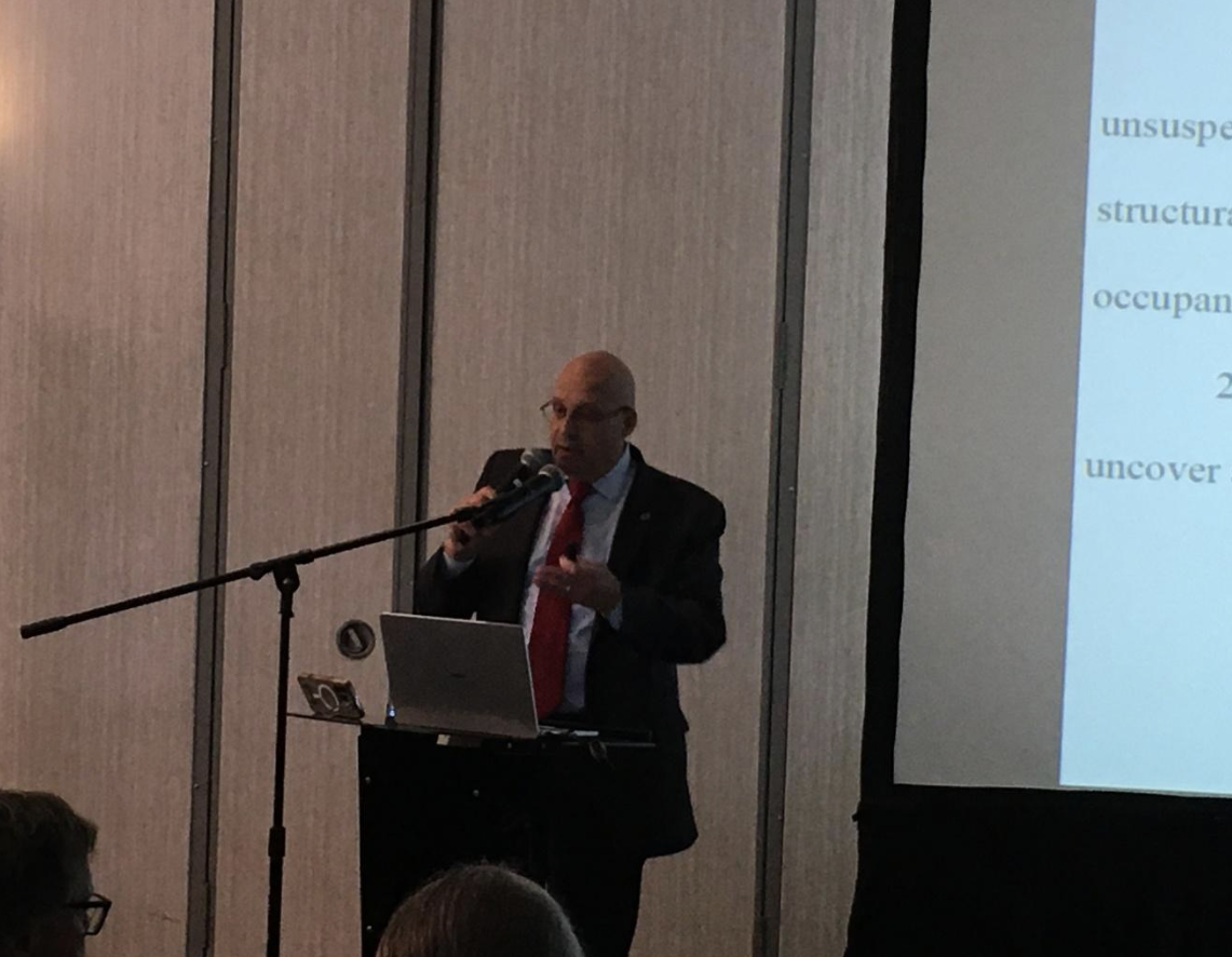 At the APRA Symposium 2026, Barry Ansbacher, in a suit and red tie, speaks into a microphone at a podium with a laptop, standing next to a projected presentation screen in a dimly lit room.