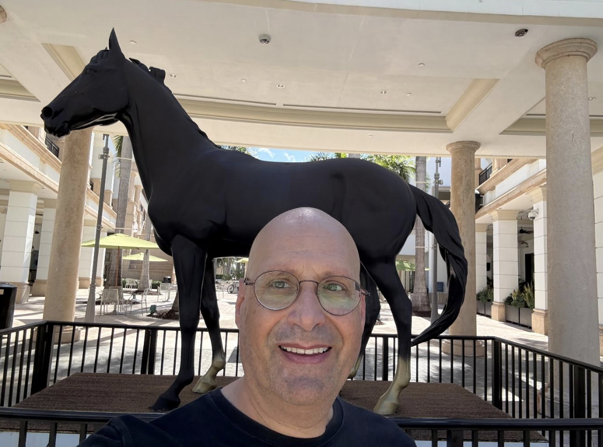 A smiling man with glasses takes a selfie in front of a large black horse statue outdoors, surrounded by a black metal fence, with columns and buildings visible—capturing the moment at the 2026 APRA Symposium.