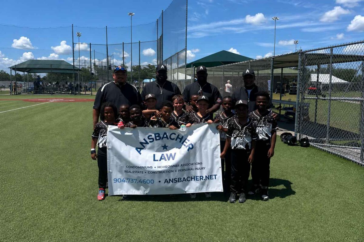 A youth baseball team called the Black Aces and four adults pose together on a sunny field, holding a white "Amsbacher Law" banner. Players wear black uniforms and hats; sports equipment and fencing are visible in the background.
