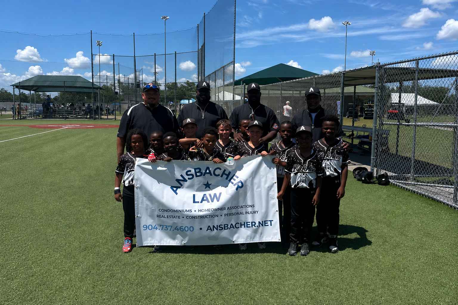 A youth baseball team called the Black Aces and four adults pose together on a sunny field, holding a white "Amsbacher Law" banner. Players wear black uniforms and hats; sports equipment and fencing are visible in the background.