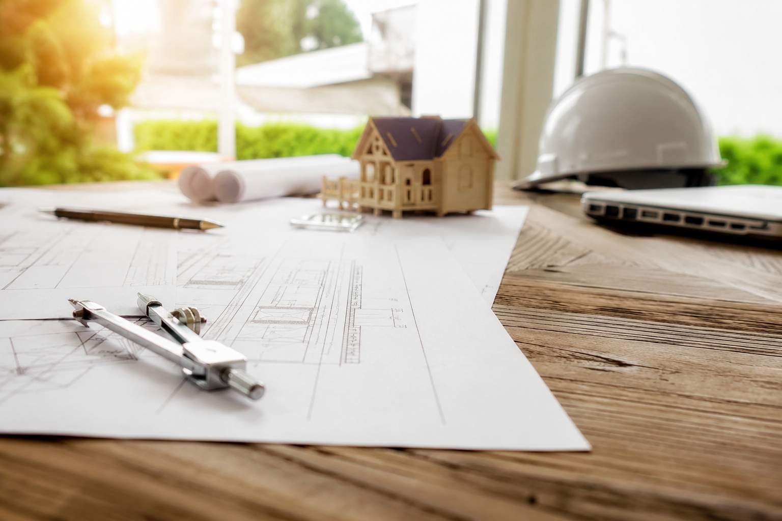A workspace with architectural blueprints, a compass, a pen, a small model house, a white safety helmet, and a laptop on a wooden desk—perfect for preparing construction contracts or planning remodeling projects with views of greenery outside.