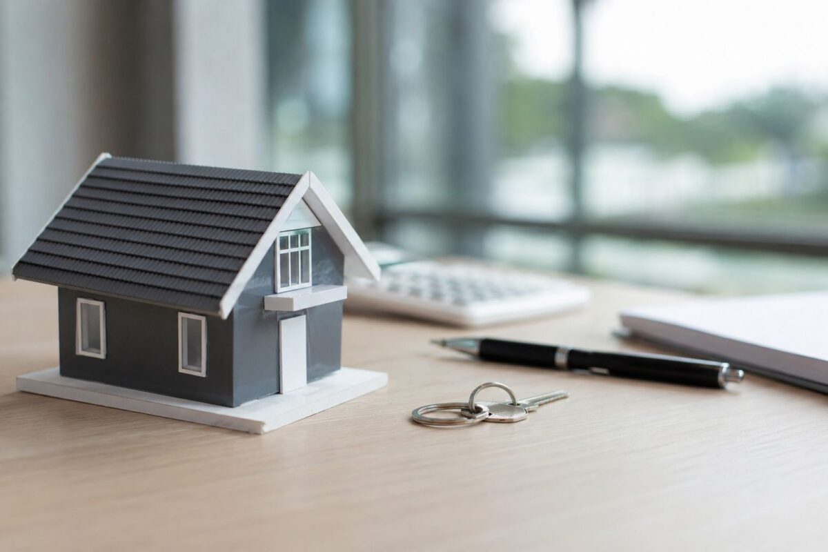 A small model house, a set of keys, a pen, a notebook, and a calculator rest on a wooden desk near a window—perfect essentials for reviewing common contingencies in your home sale contract.