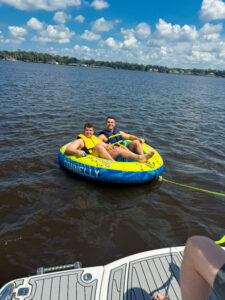 Two people wearing life jackets sit in a yellow and blue inflatable tube on a Jacksonville lake, smiling at the camera. A third person's leg is visible on a boat platform—perhaps a fellow Summer Clerk from Ansbacher Law—under a partly cloudy sky.