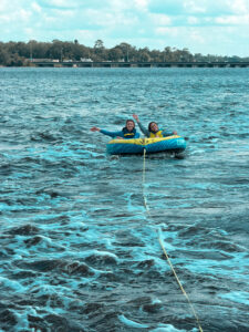 Two people ride an inflatable blue tube being towed on a large body of water near Jacksonville, smiling and waving toward the camera, with trees and a bridge in the background under a partly cloudy sky. Summer Clerks at Ansbacher Law enjoy some fun in the sun!.
