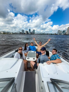 Five young adults, including Summer Clerks from Ansbacher Law, sit and smile on a boat in Jacksonville, with city buildings and a bridge in the background under a partly cloudy sky. One sits shirtless with arms raised, celebrating.