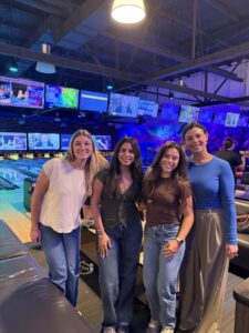 Four women, including Summer Clerks from Ansbacher Law, smiling and posing together at a Jacksonville bowling alley. TV screens and neon lights create a lively atmosphere in the background near the bowling lanes.